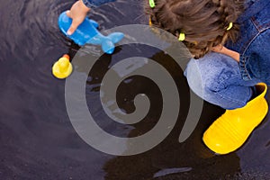 Beautiful girl in denim suit and yellow rubber boots plays with plastic duck and whale in a puddle