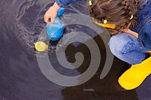 Beautiful girl in denim suit and yellow rubber boots plays with plastic duck and whale in a puddle