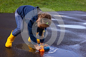 Beautiful girl in denim suit and yellow rubber boots plays with plastic duck and whale in a puddle