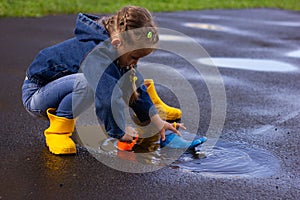 Beautiful girl in denim suit and yellow rubber boots plays with plastic duck and whale in a puddle