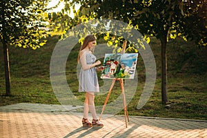 Beautiful girl draws a picture in the park using a palette with paints and a spatula. Easel and canvas with a picture. Summer is a