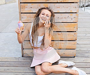 Beautiful girl with dreadlocks in pink skirt sitting on the veranda and eating colorful ice-cream cone on a warm summer evening.