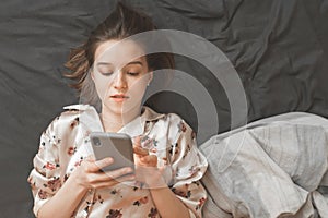 Beautiful girl dressed in pajamas, sleeping in bed and using a smartphone, top view. Woman with a smartphone in her arms is lying