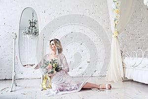 Beautiful girl dressed in peignoir and underwear sits on floor near mirror and holds bouquet in her hands.