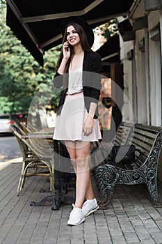 A beautiful girl dressed in a stylish white dress, a black jacket and a black hat is standing near the marble coffee table and a w