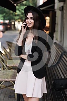 A beautiful girl dressed in a stylish white dress, a black jacket and a black hat is standing near the marble coffee table and a w