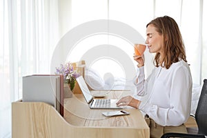Beautiful girl drinking a cup of coffee or tea in the morning with laptop computer and cell phone on wood table in bedroom