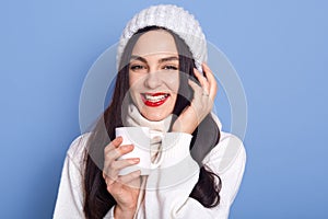 Beautiful girl enjoying cup of tea isolated on blue background, brunette female with red lips, girl wearing white sweater and cap