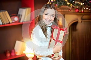 Beautiful girl in the evening in the room holds a gift wrapped in red ribbon and smiles