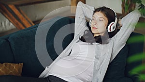 Beautiful girl expecting mother is listening to music wearing headphones and relaxing on sofa at home enjoying rest and