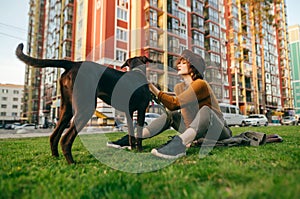 Beautiful girl in fashionable clothes and hat playing with puppy on the lawn in the yard on the background of modern colored