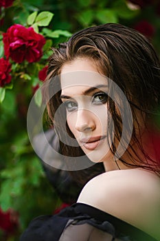 Beautiful girl in the garden with roses. Portrait of a woman with big lips in black clothes on a background of red flowers. Greens