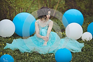 Beautiful girl graduate in a blue dress is sitting on the grass near a large blue and white balloons. Pensive elegant