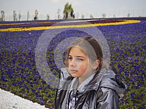 A beautiful girl in a gray jacket sits against the background of a multicolored flower meadow and looks into the distance. Young