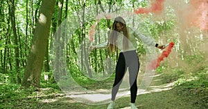Beautiful girl in a green forest spins around while holding two red smoke bombs. Slow motion