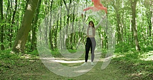 Beautiful girl in a green forest spins around while holding two red smoke bombs. Slow motion