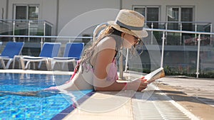 Beautiful girl in hat and sunglasses reading book at pool. Young woman relaxing at warm sunny day during vacation