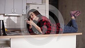 Beautiful girl in her 20`s lying on her kitchen table surface an texting someone.