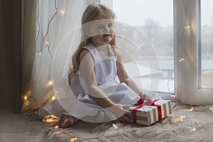 Beautiful girl holding gift sitting on a window sill. Christmas concept