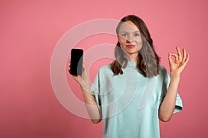 Beautiful girl holds mobile phone and makes okay gesture on a pink background. Emotions and pleasant feelings concept.