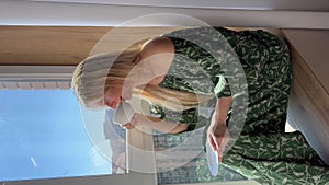 A beautiful girl in home clothes sits near the window and drinks coffee or tea from a white cup.