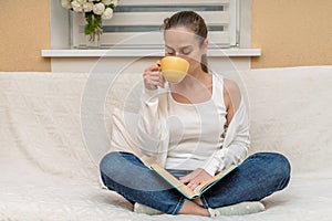 Beautiful girl at home sitting on the sofa, reading a book and arranging a coffee break, relaxation concept, good at home