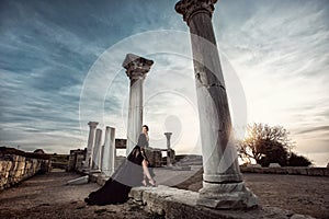 A beautiful girl in a long black dress poses at sunset amid the sea, antique columns and ruins.