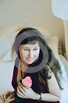 Beautiful girl with long dark hair in a burgundy dress sits on the bed and holds a rose in her hands