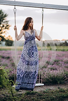 Beautiful girl in a long dress on a swing in a lavender field at sunset. Soft focus