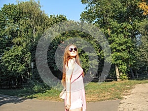 Beautiful girl with long golden hair in sunglasses looks at the sun. Young woman in pink clothes walking in a park on a clear