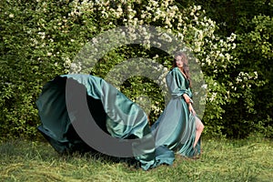 A beautiful girl in a long green silk dress on the background of a flowering tree. The hem of the dress develops in the wind . Cop