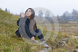 A beautiful girl with long hair in black clothes sits on the grass on the outskirts of the city and looks to the side