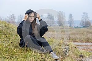 A beautiful girl with long hair in black clothes sits on the grass on the outskirts of the city. She puts a hood over her head and