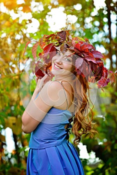 Beautiful girl with long hair in blue dress, in a wreath of red and green leaves standing against bright fall bushes