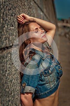 Beautiful girl with long hair and in a denim jacket stands near the wall in the summer.