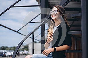 Beautiful girl with long hair and glasses sitting on metal stairs on the wooden background of house with vertical boards. Woman