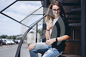 Beautiful girl with long hair and glasses sitting on metal stairs on the wooden background of house with vertical boards. Woman