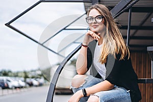 Beautiful girl with long hair and glasses sitting on metal stairs on the wooden background of house with vertical boards. Woman