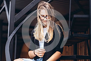 Beautiful girl with long hair and glasses sitting on metal stairs on the wooden background of house with vertical boards. Woman