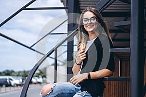 Beautiful girl with long hair and glasses sitting on metal stairs on the wooden background of house with vertical boards. Woman