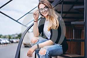Beautiful girl with long hair and glasses sitting on metal stairs on the wooden background of house with vertical boards. Woman