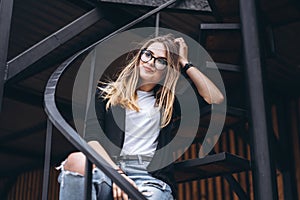 Beautiful girl with long hair and glasses sitting on metal stairs on the wooden background of house with vertical boards. Woman