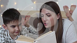 A beautiful girl with long hair reads a book to a small boy lying on a bed and kisses him having a good mood.HD