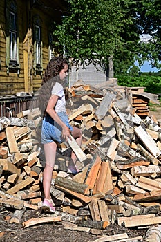 beautiful girl with long hair in white T-shirt and denim shorts puts firewood in pile. Concept of harvesting firewood for winter.