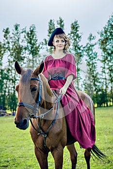 Beautiful girl in a long red dress and in a black hat with a cocked hat riding a brown horse