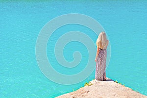 Beautiful girl with long white hair in a long dress standing on the shore of the lake with blue water in a Sunny bright day