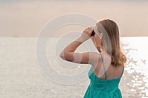 Girl looking in binoculars at sea on twilight. Young woman in turquoise dress observes seascape in cloud weather. Hope and sadness