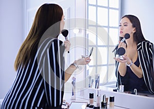 Beautiful girl looking in the mirror and applying cosmetic with a big brush