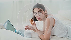 Beautiful girl lying on the bed in morning, using laptop. Young woman eating croissant, typing, browsing the Internet.