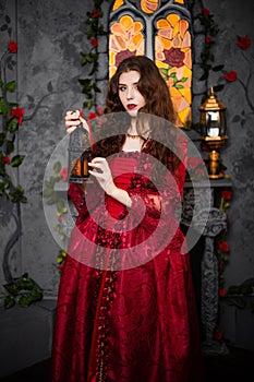 A beautiful girl in a magnificent red dress of the Rococo era stands against a fireplace, a window and flowers with a lamp with ca